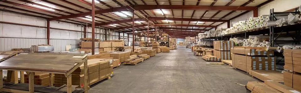 A large warehouse interior with rows of boxed flooring and pallets on a concrete floor; no people are visible.