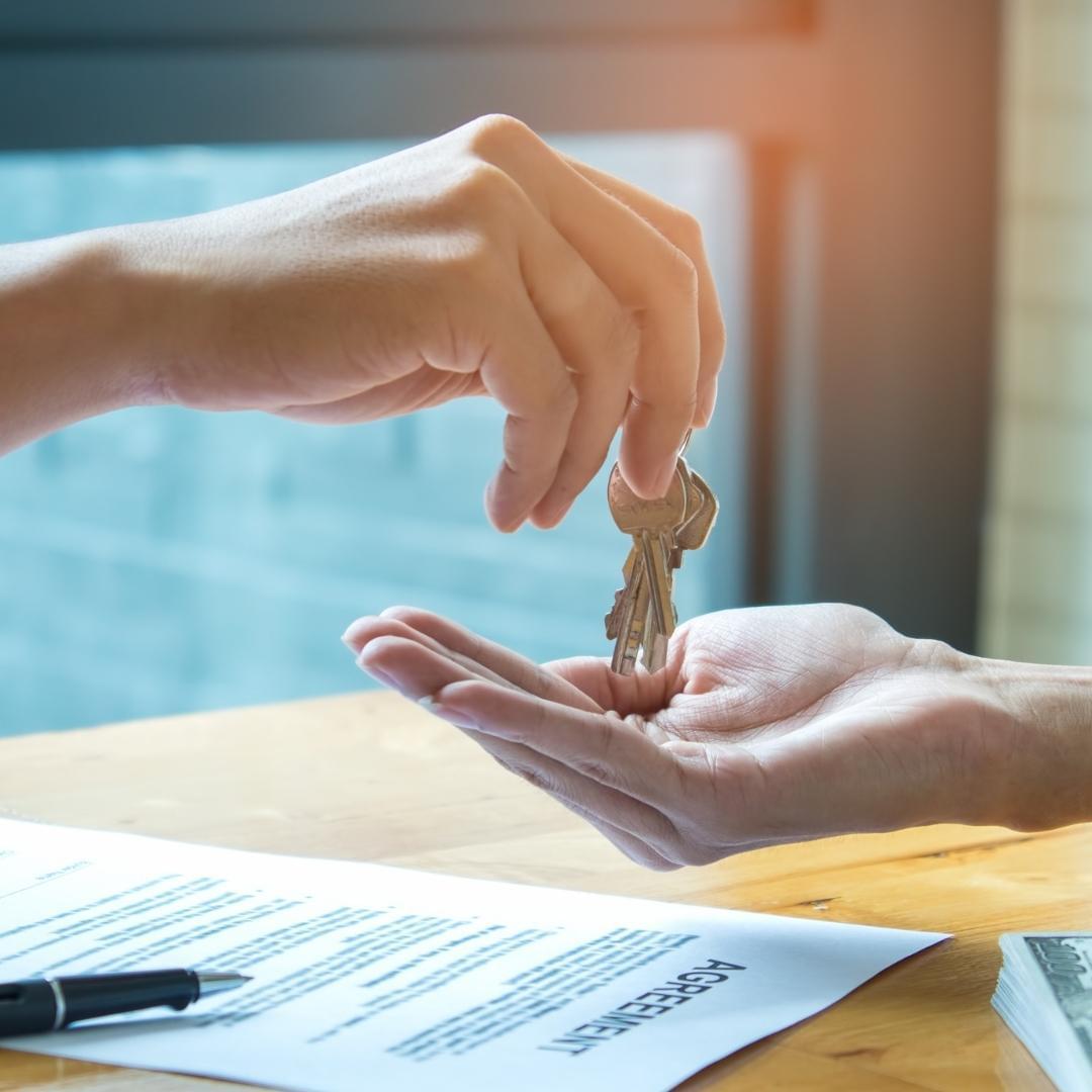 Two people exchange metal keys above a printed document on a wooden desk.